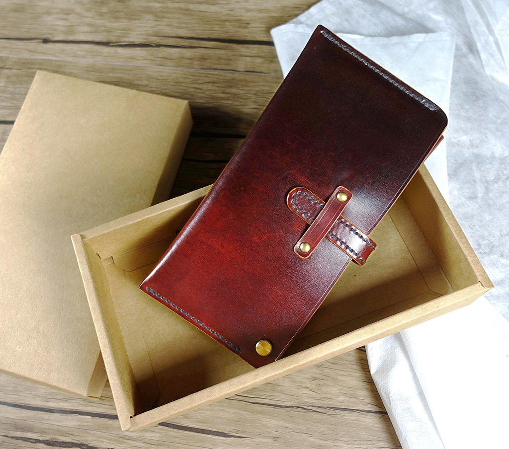翻轉長夾成品平放於紙盒中,呈現皮革自然色澤與手縫邊線細節,螺絲扣轉軸設計清晰可見。