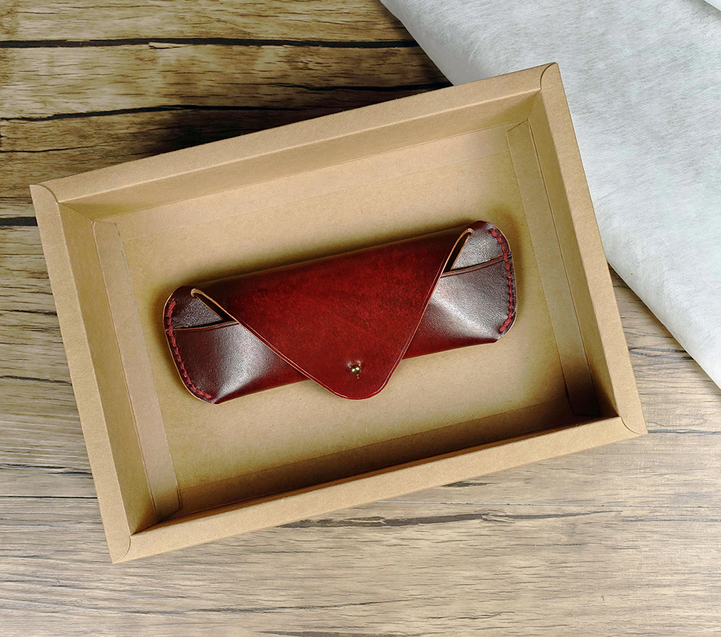 手作皮革眼鏡皮套置於牛皮紙禮盒中展示,紅棕色染色效果明顯,翻蓋式設計與金屬釦收合細節清楚呈現。