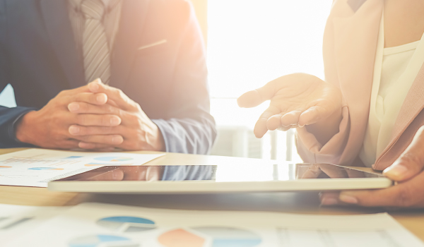 Two business professionals in formal attire discussing documents and data charts during a meeting, with a digital tablet on the table.
