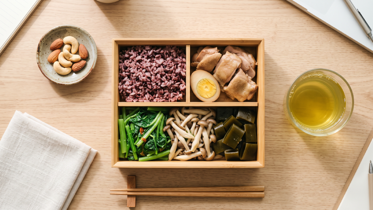 俯拍一張木質餐桌上的台灣外食穩糖午餐組合。一個格狀便當盒內裝著去皮滷雞腿、三格滿滿的深綠色燙青菜與炒鴻喜菇、半碗紫米五穀飯；旁邊擺著一杯無糖綠茶與一小碟（約 6 顆）原味低溫烘焙腰果與杏仁。